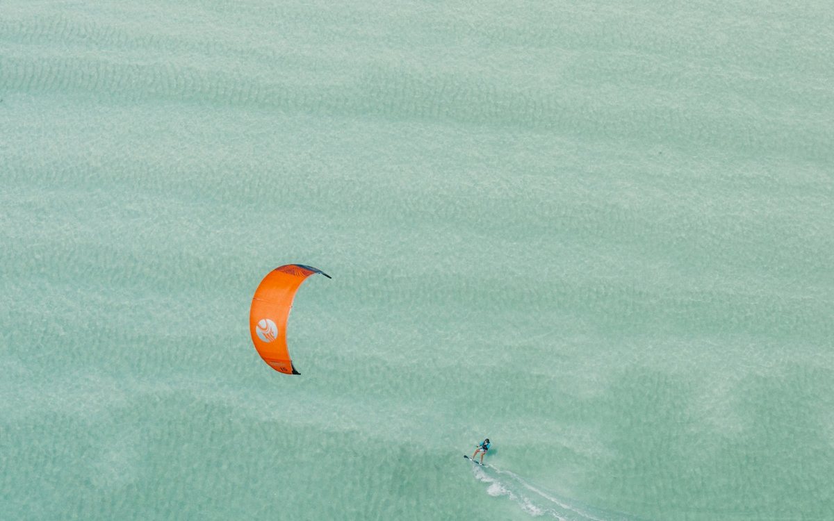 A kitesurfer with an orange kite gliding over shallow turquoise water
